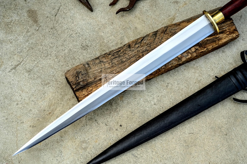 May include: A silver and gold dagger with a wooden handle rests on a wooden plank. A black leather sheath is positioned to the right of the dagger. The dagger has a sharp, pointed blade and a gold-colored guard.