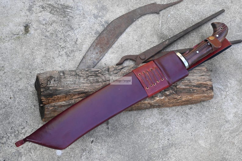 May include: A handmade knife with a dark wooden handle and a burgundy leather sheath. The sheath is stitched with brown thread. A curved blade and a pair of pliers are in the background, resting on a weathered wooden block.