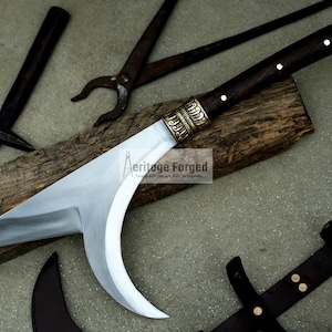 May include: A curved, silver-colored blade with a dark wood handle and brass accents. The blade is resting on a piece of wood with a hammer and pliers in the background. The text "Heritage Forged" is visible on the wood.