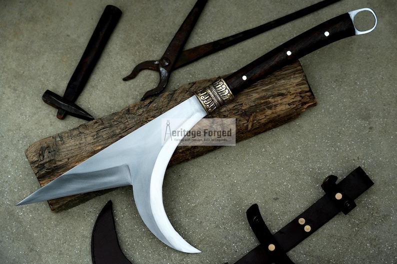 May include: A curved, silver-colored blade with a dark wood handle and brass accents. The blade is resting on a piece of wood with a hammer and pliers in the background. The text "Heritage Forged" is visible on the wood.