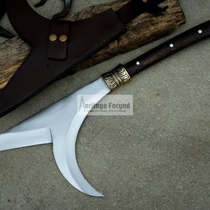 May include: A curved, silver-colored blade with a brown wooden handle and a brass accent. The blade is attached to a brown leather sheath. The text "Heritage Forged" is visible on the blade.