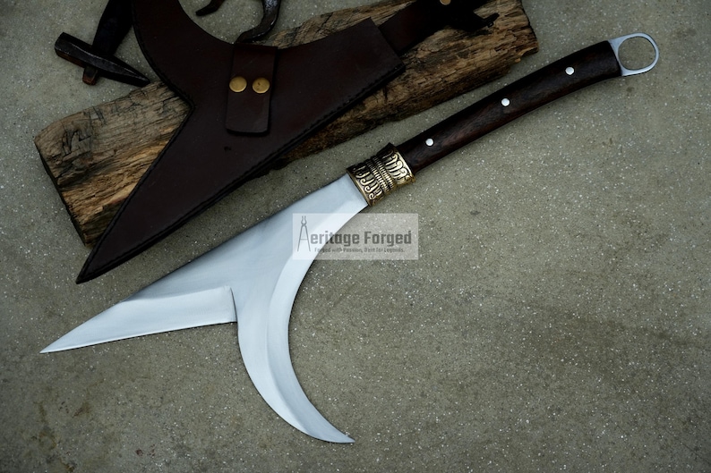May include: A curved, silver-colored blade with a brown wooden handle and a brass accent. The blade is attached to a brown leather sheath. The text "Heritage Forged" is visible on the blade.