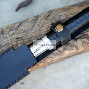 May include: Close-up of a black sword in a black leather sheath. The sword handle is black with a leather strap and a gold-coloured button. The sheath is stitched with a silver metal band.