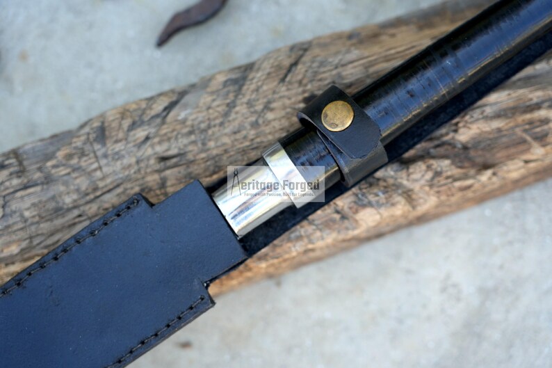 May include: Close-up of a black sword in a black leather sheath. The sword handle is black with a leather strap and a gold-coloured button. The sheath is stitched with a silver metal band.