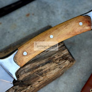 May include: A close-up of a knife with a wooden handle. The handle is made of light brown wood and has three metal rivets. The blade is made of stainless steel and has a sharp point. The knife is resting on a piece of wood.