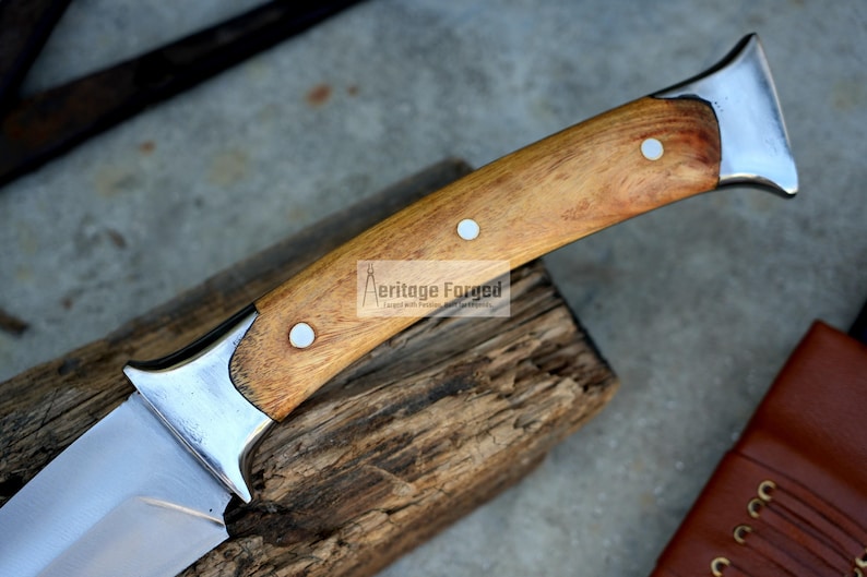 May include: A close-up of a knife with a wooden handle. The handle is made of light brown wood and has three metal rivets. The blade is made of stainless steel and has a sharp point. The knife is resting on a piece of wood.