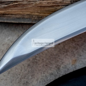 May include: Close-up of the blade of a curved knife. The blade is made of polished steel and has a sharp point. The blade is labeled "Heritage Forged" in white text.