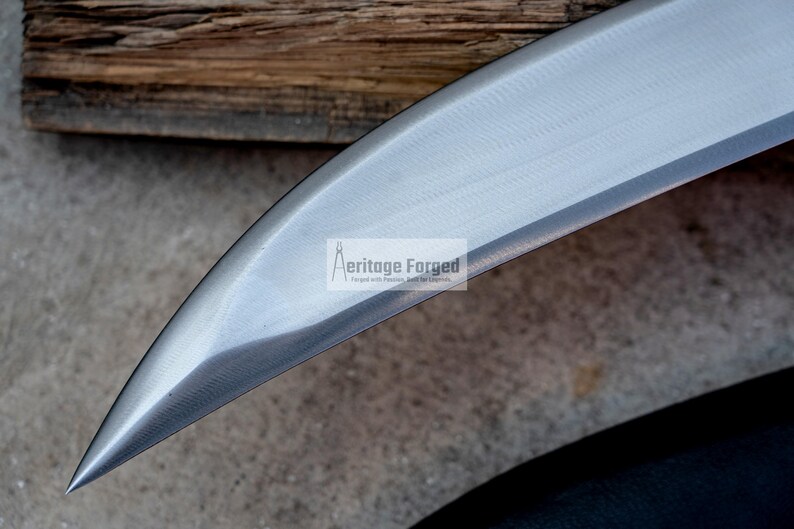 May include: Close-up of the blade of a curved knife. The blade is made of polished steel and has a sharp point. The blade is labeled "Heritage Forged" in white text.