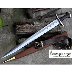 May include: A silver sword with a brown leather scabbard and a brown leather belt with a brass buckle. The sword has a wooden handle with a dark brown finish. The sword is on a gray surface with a piece of wood and some tools.