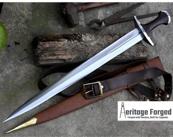 May include: A silver sword with a brown leather scabbard and a brown leather belt with a brass buckle. The sword has a wooden handle with a dark brown finish. The sword is on a gray surface with a piece of wood and some tools.