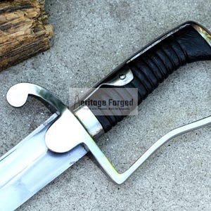 May include: Close-up of a saber with a silver blade and guard, and a black leather-wrapped handle. The text "Heritage Forged" is visible on the blade. The saber is on a gray surface.