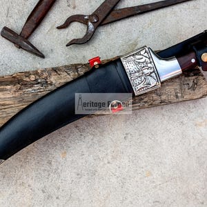 May include: A black leather-wrapped curved knife with a silver handle and a wooden sheath. The knife has intricate silver designs on the handle and blade. The knife is resting on a piece of wood.