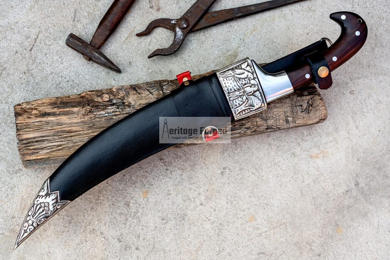 May include: A black leather-wrapped curved knife with a silver handle and a wooden sheath. The knife has intricate silver designs on the handle and blade. The knife is resting on a piece of wood.