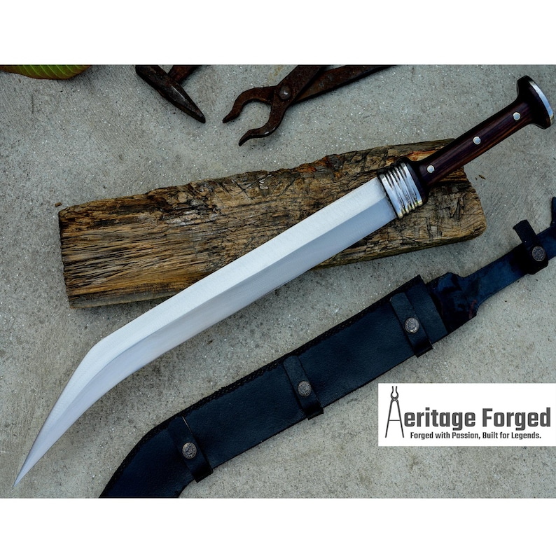 May include: A silver blade with a brown wooden handle and a black leather sheath. The blade is curved and has a sharp point. The handle is decorated with silver rings. The sheath has a metal buckle. The text "Heritage Forged" is visible in the lower right corner of the image. "Forged with Passion, Built for Legends."