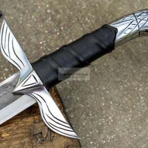 May include: Close-up of a silver sword with a black leather-wrapped handle. The blade is silver and the handle has intricate designs. The sword rests on a wooden surface, showcasing its detailed craftsmanship.