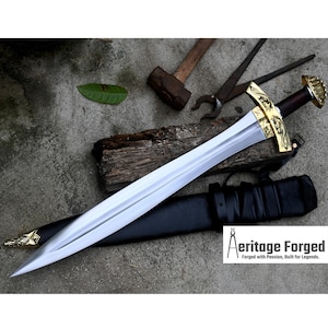 May include: A silver sword with a gold hilt and a black leather scabbard. The sword has a decorative design on the hilt and a cross-shaped pommel. The scabbard has a strap and a buckle. The sword is lying on a wooden surface with a hammer and pliers in the background. The image includes the text "Heritage Forged, Forged with Passion, Built for Legends."