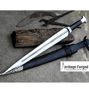 May include: A silver and black sword with a wooden handle and a leather scabbard. The sword is lying on a wooden surface with a metal tool and a piece of wood behind it. The text "Heritage Forged" is visible in the lower right corner of the image. "Forged with Passion, Built for Legends."