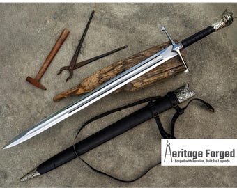 May include: A large, silver sword with a black handle and crossguard, resting on a piece of wood. The sword is in a black sheath with a strap. A hammer, tongs, and other tools are nearby. The text "Aeritage Forged" is visible.
