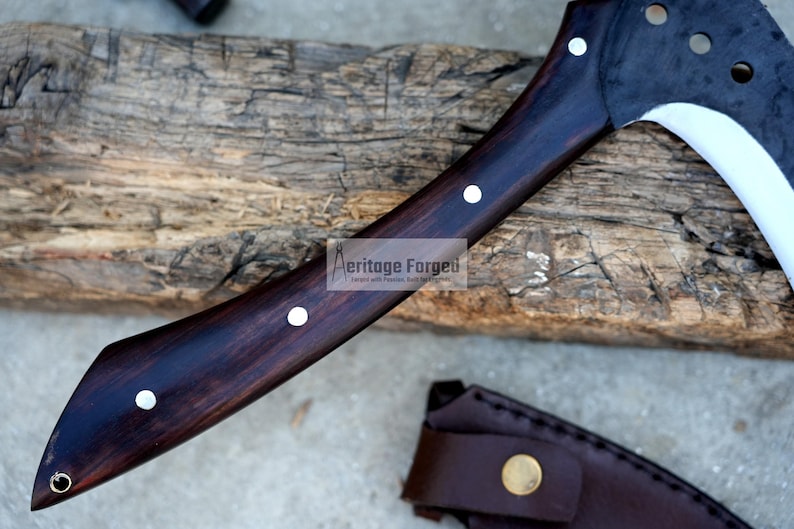 Op de afbeelding: Een donkerbruine houten handgreep met zilveren accenten en een gebogen, zilveren blad. Het blad heeft een scherpe punt en een licht gebogen rand. De handgreep is met een metalen klinknagel aan het blad bevestigd. Het blad is gedeeltelijk zichtbaar in de afbeelding. De handgreep rust op een houten oppervlak. De tekst "Heritage Forged" is zichtbaar op de handgreep.