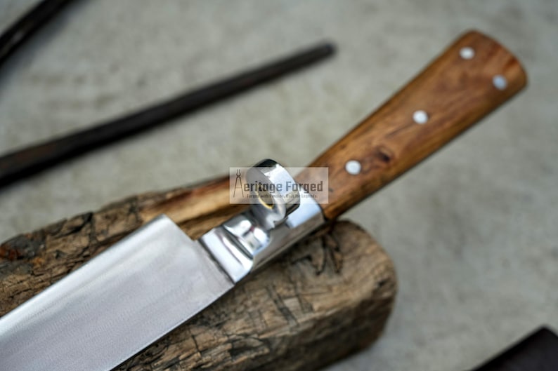 May include: A stainless steel knife with a wooden handle. The handle has a ring on the top and is made of dark wood with white accents. The knife is resting on a piece of wood.