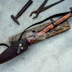 K&ouml;nnte beinhalten: Ein braunes Lederscheide mit einem Messer mit Holzgriff und einem braunen Lederriemen. Das Messer hat einen braunen Holzgriff mit einem Messingakzent. Das Messer liegt auf einer Holzoberfl&auml;che.