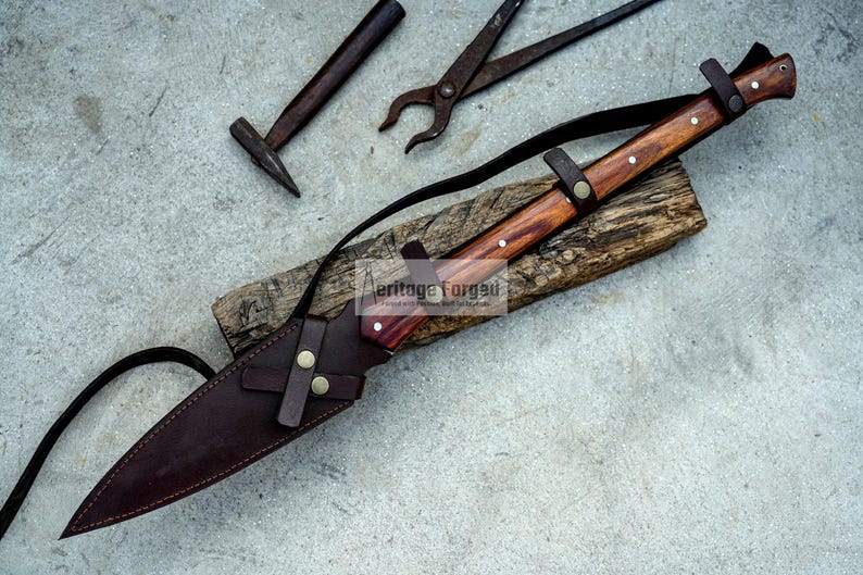 K&ouml;nnte beinhalten: Ein braunes Lederscheide mit einem Messer mit Holzgriff und einem braunen Lederriemen. Das Messer hat einen braunen Holzgriff mit einem Messingakzent. Das Messer liegt auf einer Holzoberfl&auml;che.