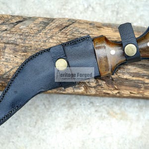 May include: A black leather sheath with a curved blade and a wooden handle. The handle has a brown wood grain and a brass accent. The sheath has a brass button and the text "Heritage Forged" is printed on the sheath.