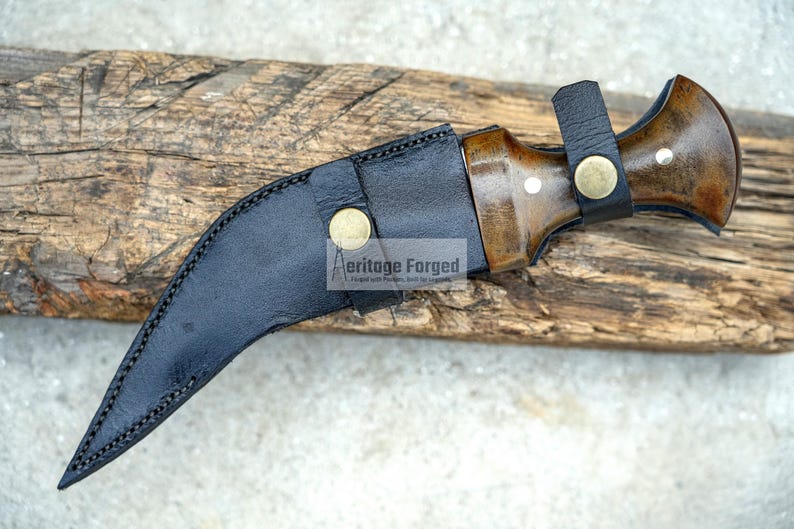 May include: A black leather sheath with a curved blade and a wooden handle. The handle has a brown wood grain and a brass accent. The sheath has a brass button and the text "Heritage Forged" is printed on the sheath.
