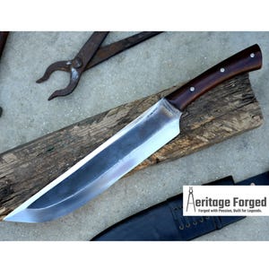 May include: A long, silver blade with a brown wooden handle. The blade is sharp and has a slight curve. The handle has a silver metal accent. The knife is resting on a wooden surface. The text "Heritage Forged" is visible in the lower right corner of the image. "Forged with Passion, Built for Legends."
