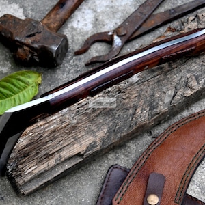 May include: A handcrafted knife with a dark wooden handle and a silver blade rests on a weathered wooden plank. A brown leather sheath, hammer, and pliers are also visible. A green leaf adds a natural element to the composition.