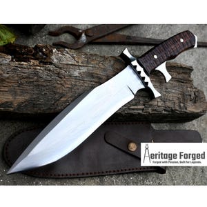 Puede incluir: Un cuchillo grande de hoja fija plateado con un mango marr&oacute;n texturizado. El cuchillo tiene una funda de cuero con un bot&oacute;n de metal. El texto "Heritage Forged" es visible en la funda. "Forjado con pasi&oacute;n, construido para leyendas."