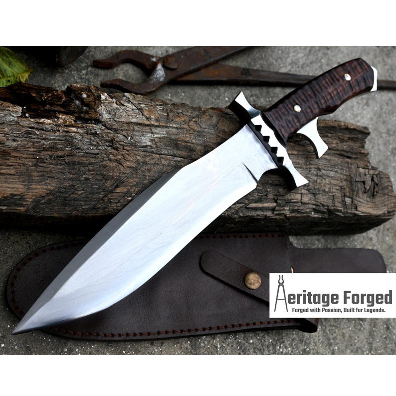 Puede incluir: Un cuchillo grande de hoja fija plateado con un mango marr&oacute;n texturizado. El cuchillo tiene una funda de cuero con un bot&oacute;n de metal. El texto "Heritage Forged" es visible en la funda. "Forjado con pasi&oacute;n, construido para leyendas."