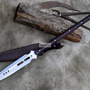 May include: A long, silver-colored spear with a brown wooden handle and a leather sheath. The spear has a pointed tip and a decorative design on the blade.