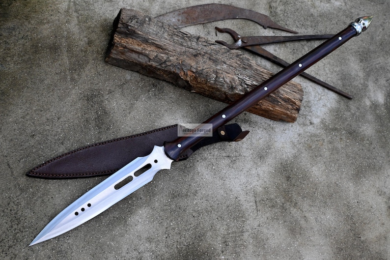 May include: A long, silver-colored spear with a brown wooden handle and a leather sheath. The spear has a pointed tip and a decorative design on the blade.