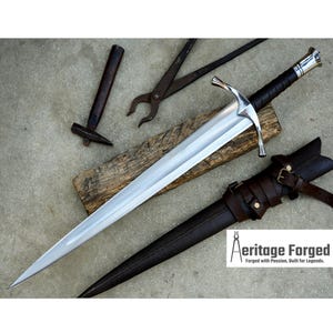Pode incluir: Uma espada prateada com um cabo envolto em couro e uma bainha de couro marrom. A espada está sobre uma superfície de madeira com um martelo e alicates por perto. O texto "Heritage Forged" é visível no canto inferior direito da imagem.