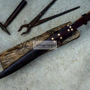 May include: A black leather knife sheath with brass rivets and a wooden handle. The sheath is resting on a piece of wood with the text "Heritage Forged" printed on it. The knife is not visible in the image.