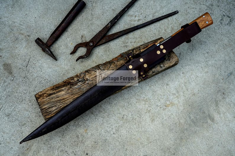 May include: A black leather knife sheath with brass rivets and a wooden handle. The sheath is resting on a piece of wood with the text "Heritage Forged" printed on it. The knife is not visible in the image.
