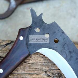 Op de afbeelding: Een gebogen, zwart metalen blad met een houten handvat. Het blad heeft een zilveren rand en verschillende gaten. Het handvat is donkerbruin met witte accenten. Het blad is gelabeld "Heritage Forged".