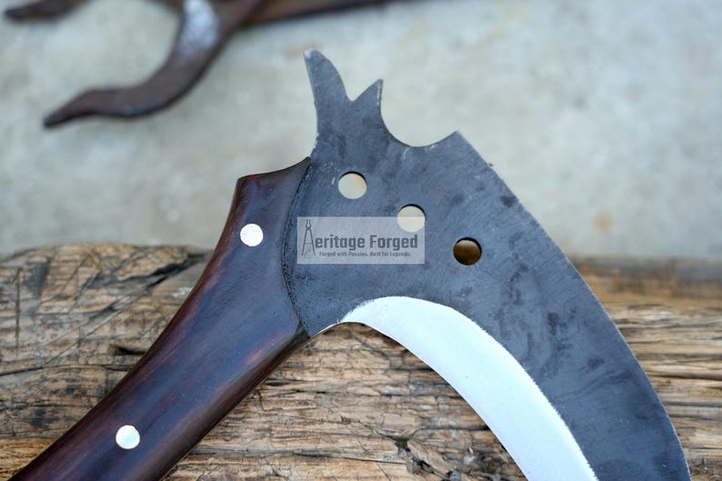 Op de afbeelding: Een gebogen, zwart metalen blad met een houten handvat. Het blad heeft een zilveren rand en verschillende gaten. Het handvat is donkerbruin met witte accenten. Het blad is gelabeld "Heritage Forged".