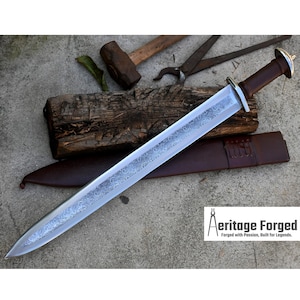 Può includere: Una spada d'argento con un designo inciso dettagliato sulla lama. La spada ha un'impugnatura in pelle marrone e poggia su una superficie di legno con un fodero in pelle marrone. Il testo "Heritage Forged, Forged with Passion, Built for Legends." è visibile nell'angolo in basso a destra dell'immagine.