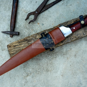 May include: A brown leather-sheathed knife with a silver blade and a wooden handle. The knife is on a piece of wood with the text "Heritage Forged" on the sheath.