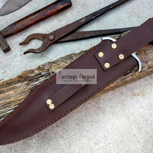 Op de afbeelding: Een bruine leren schede met messing accenten voor een mes. De schede ligt op een stuk hout met de tekst "Heritage Forged" erop gedrukt.