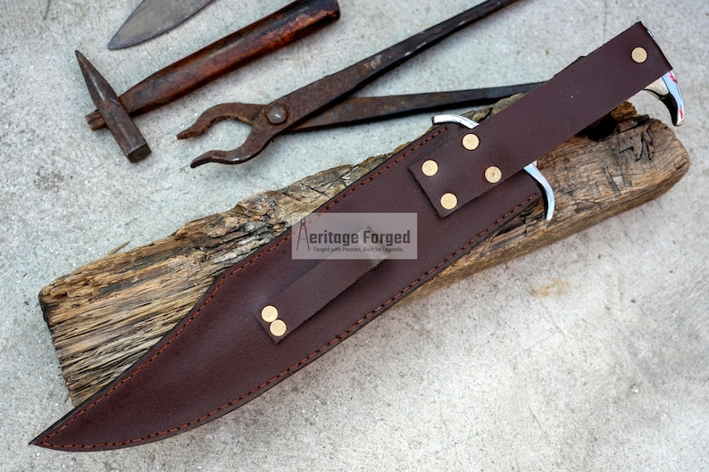 Op de afbeelding: Een bruine leren schede met messing accenten voor een mes. De schede ligt op een stuk hout met de tekst "Heritage Forged" erop gedrukt.
