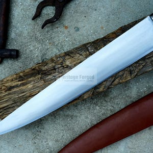 May include: A long, silver blade with a wooden handle and a brown leather sheath. The blade is sharp and has a slight curve. The sheath has a strap with a buckle. The text "Heritage Forged" is visible on the blade.