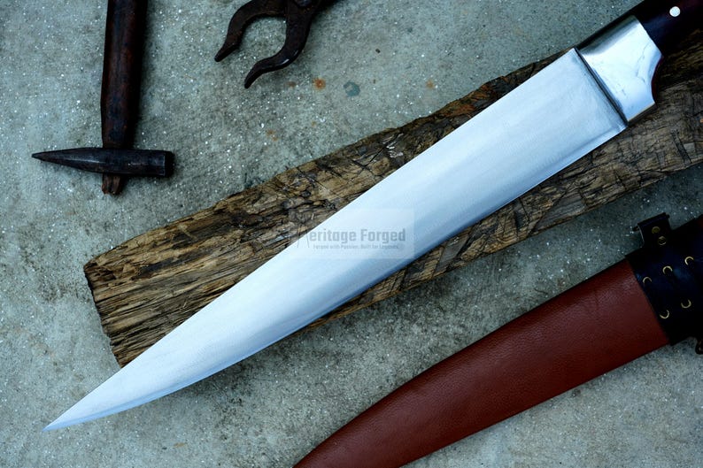 May include: A long, silver blade with a wooden handle and a brown leather sheath. The blade is sharp and has a slight curve. The sheath has a strap with a buckle. The text "Heritage Forged" is visible on the blade.