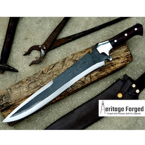 Op de afbeelding: Een zwart en zilveren mes met een houten handvat. Het mes is scherp en heeft een gekarteld lemmet. Het mes ligt op een houten oppervlak. Het mes wordt geleverd met een leren schede. De tekst "Heritage Forged" is zichtbaar in de afbeelding.  Gesmeed met passie, gebouwd voor legendes.