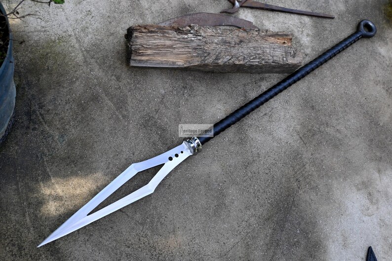 Puede incluir: Una lanza negra y plateada con una hoja en forma de diamante y un mango negro. La lanza mide aproximadamente 1,5 metros de largo y tiene un anillo en la parte superior del mango.