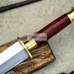 May include: A close-up of a handcrafted sword with a polished steel blade and a dark wood handle. The handle is capped with gold-colored metal. The sword rests on a wooden surface. The text "Heritage Forged" is visible.
