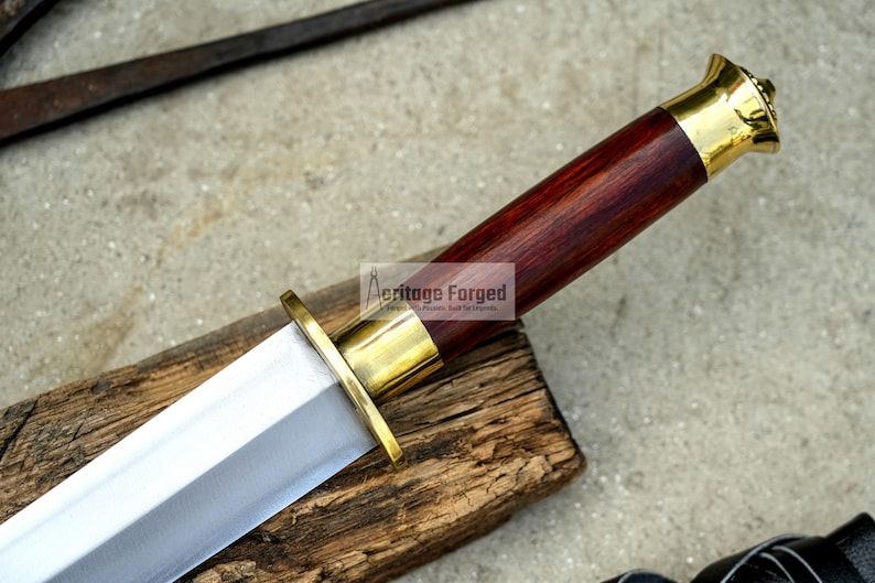 May include: A close-up of a handcrafted sword with a polished steel blade and a dark wood handle. The handle is capped with gold-colored metal. The sword rests on a wooden surface. The text "Heritage Forged" is visible.