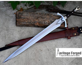 May include: A silver sword with a brown leather scabbard. The sword has a decorative handle with a pointed tip. The scabbard has a leather strap with a metal buckle. The sword is on a wooden surface. The text "Heritage Forged" is visible in the lower right corner of the image. "Forged with Passion, Built for Legends."